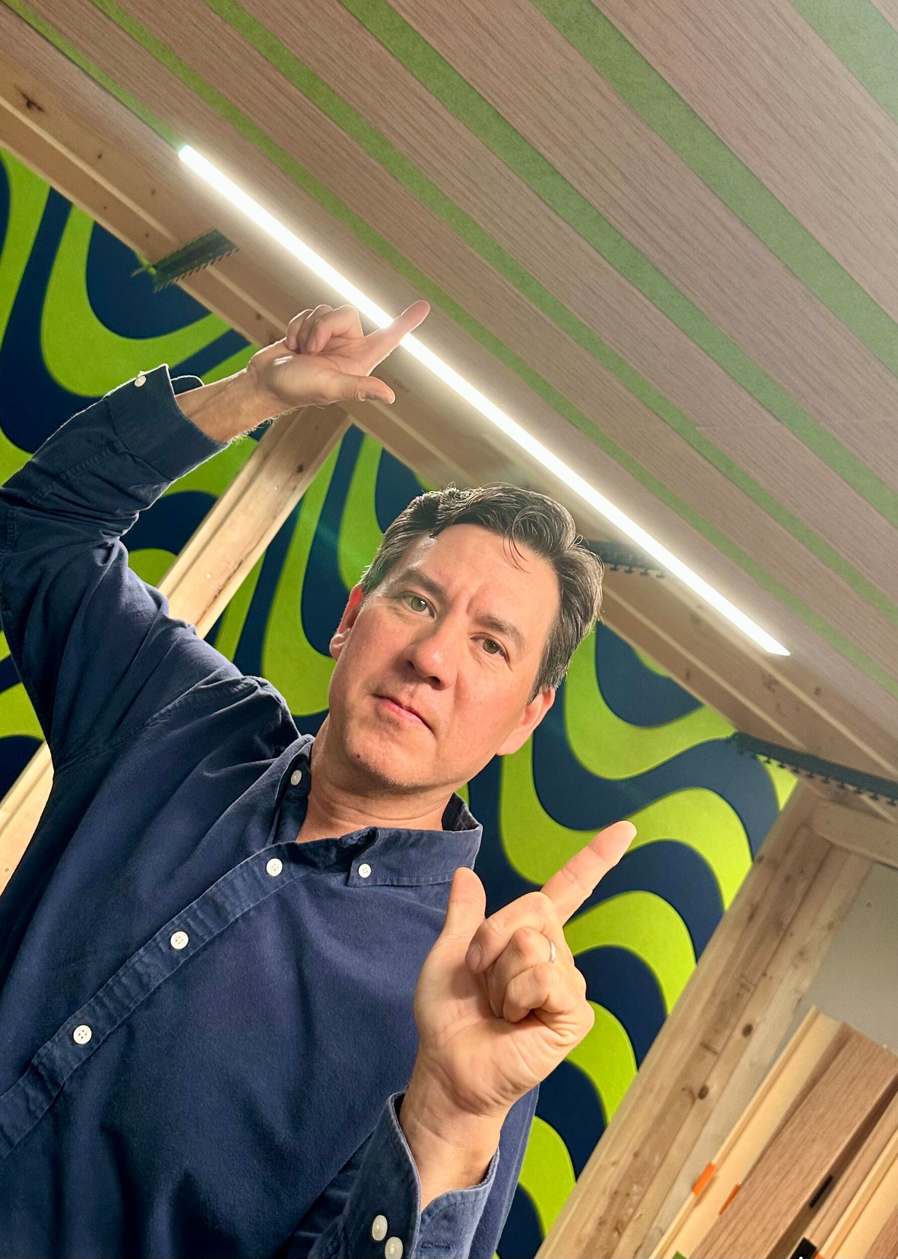Man in a Blue Shirt Pointing Upward in a Room with Wooden Beams and Patterned Wall